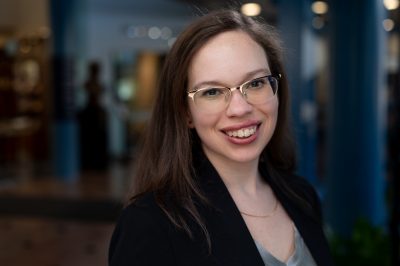 A woman with glasses with gold frames and straight, brown hair smiles at the camera.