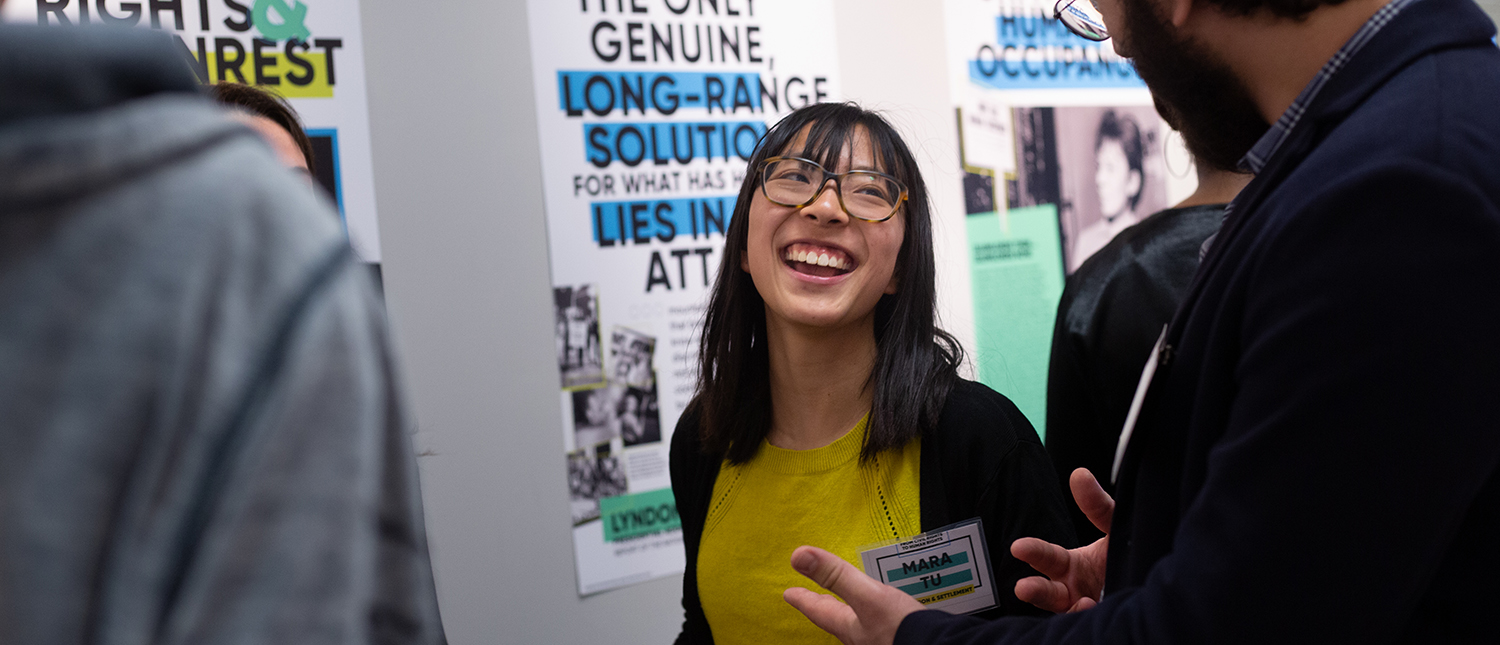 A woman in a yellow shirt and glasses is laughing during a poster session.