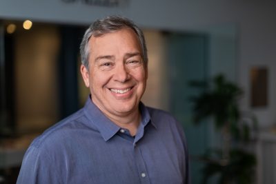 A man in an indigo button down shirt smiles at the camera.