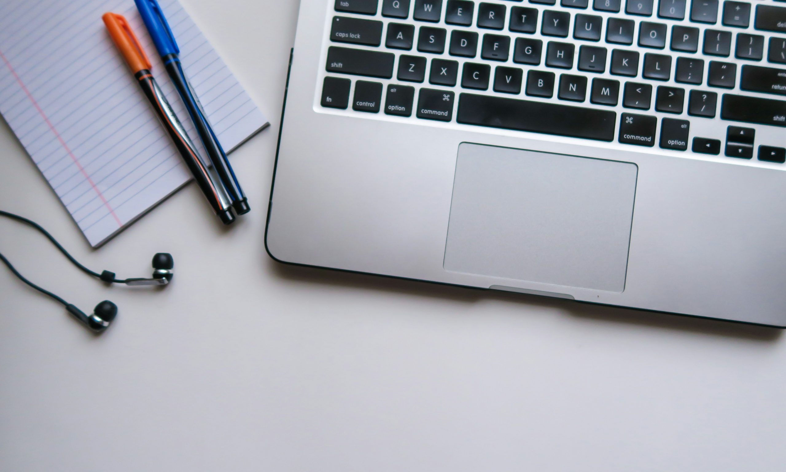 The keyboard of a laptop, a pad of paper, two pens and a pair of headphones. 