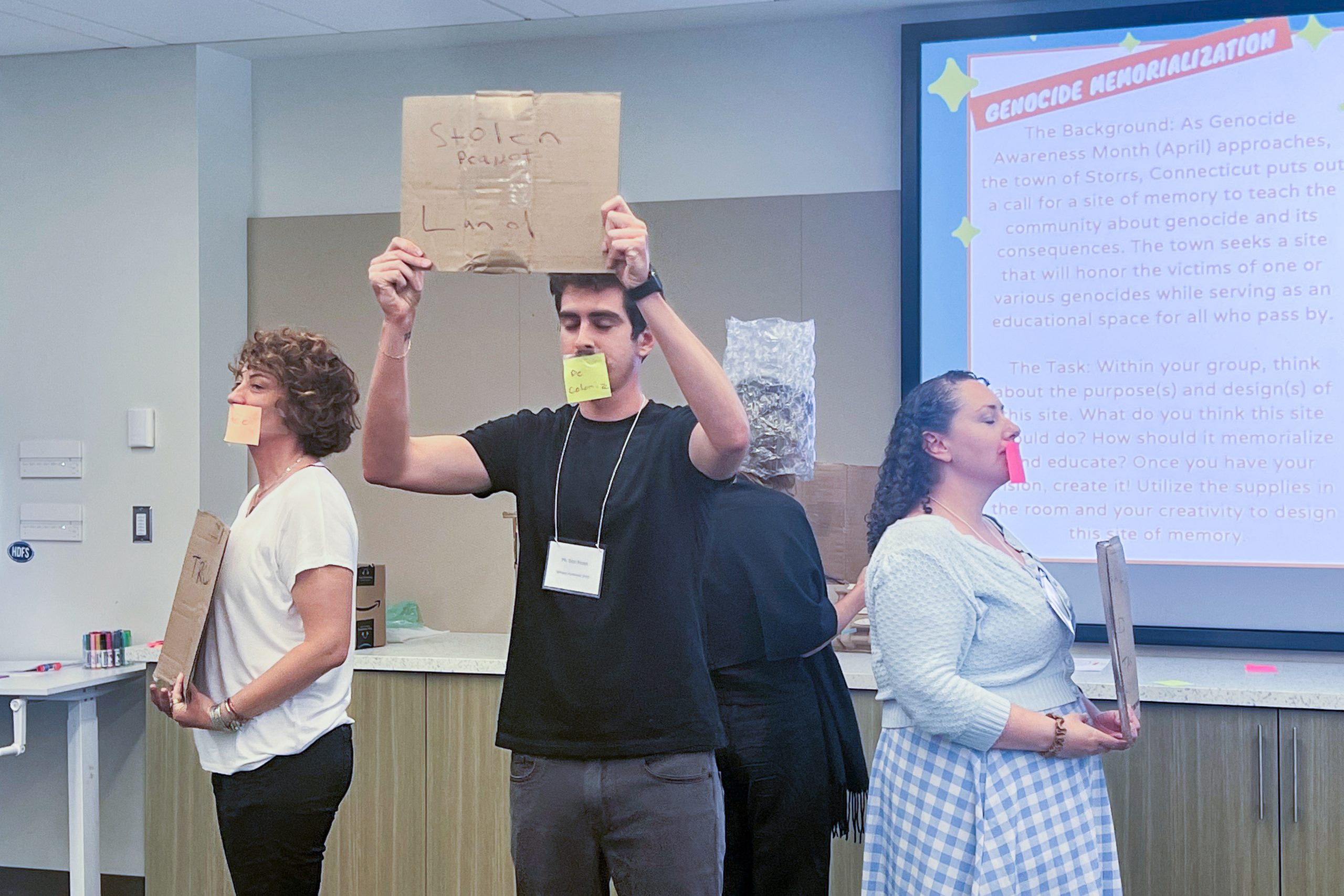 Four people stand in front of a seminar presentation in a circle with their backs to one another. The slide behind then is titled "Genocide Memorialization." They have their eyes close, a sticky note over their mouths, and are holding cardboard signs. One has what looks like bubble wrap on their head. The person facing the camera is holding their sign up and it reads "Stolen Land."