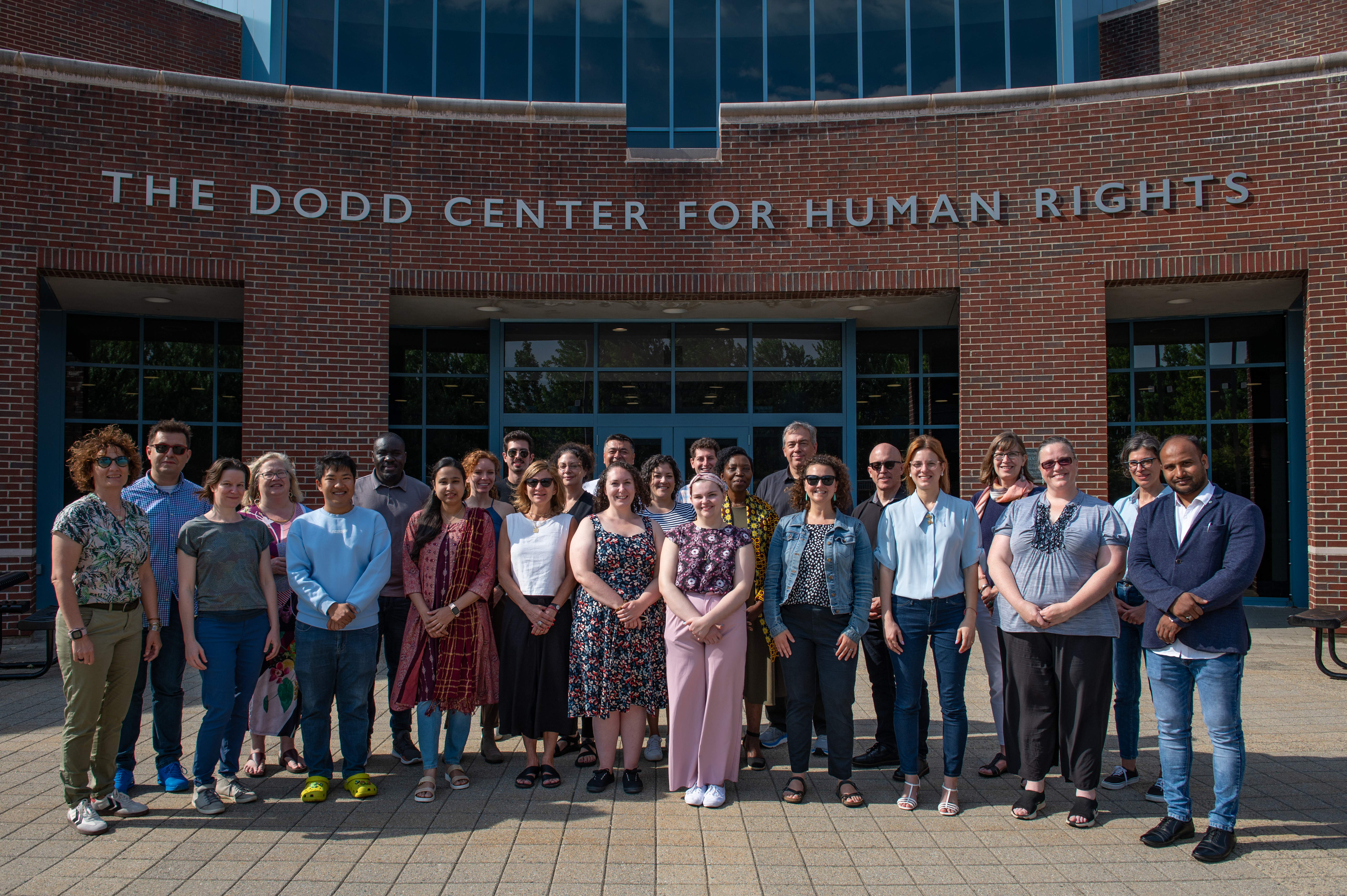 A group of staff and faculty members from the Gladstein Family Human Rights Institute stand together in two rows for a group photo in front of The Dodd Center for Human Rights.