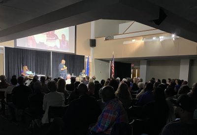 A person stands lit on a stage giving a presentation amongst a seated crowd. 