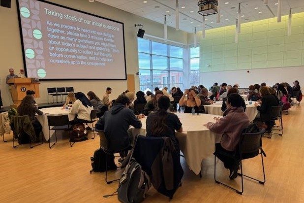 A seminar of people listening to a lecturer. The slide on the presentation reads "Taking stock of our initial questions."