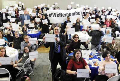 A group of people are gathered in a conference room. Everyone is holding a piece of printer paper with something typed on it. In the back of the room, people hold a banner that reads "#WeRemember."